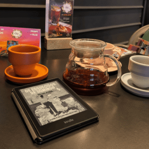 A Kindle displaying Forgotten on Sunday by Valérie Perrin on a café table beside a cup of tea and a glass coffee maker.